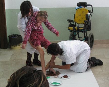 Immagine di un alunna disabile che disegna con i piedi con l'aiuto dell'assistente e dell'insegnante